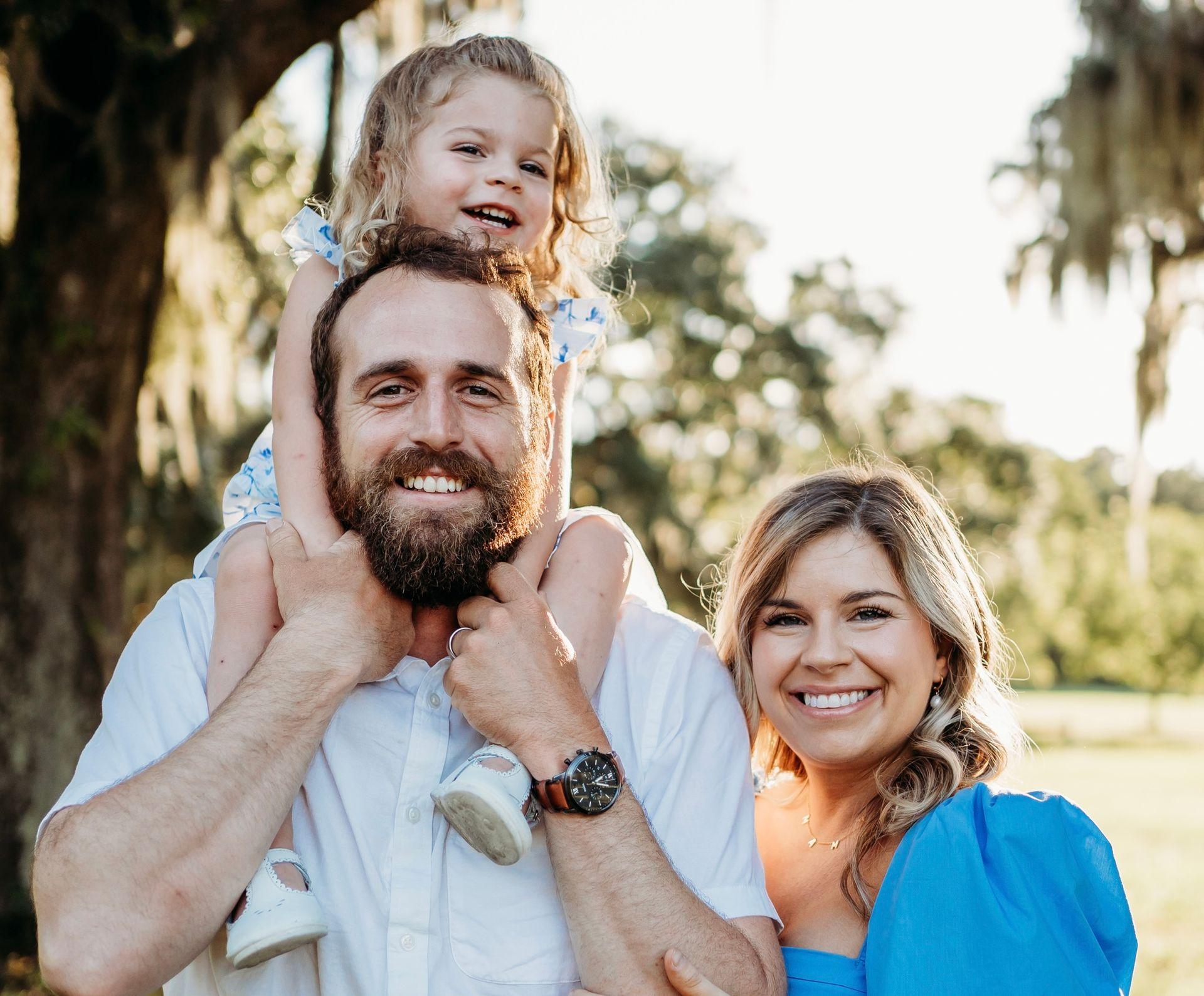 Smiling family outdoors, man holding child on shoulders next to woman in blue dress.