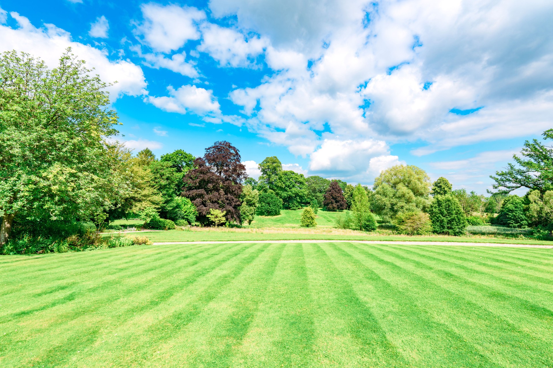 Vibrant green lawn stripes leading to lush trees in cotswolds park under a cloudy blue sky